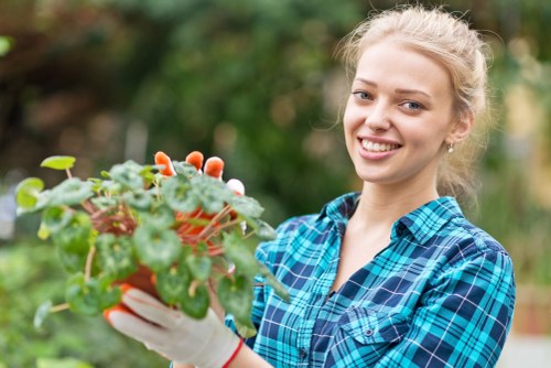 A young woman in a blue plaid shirt and white gardening gloves is smiling while holding a potted plant with green and reddish leaves. She is standing outdoors in a garden or backyard setting, with blurred trees and shrubbery in the background. The scene suggests a pleasant day of gardening or plant care, with natural lighting highlighting her cheerful expression and the healthy foliage of the plant, which appears to be a type of decorative or flowering shrub. The environment features a lush, green landscape typical of garden spaces in Balham, London, and the woman's attire and the plant she holds reflect professional gardening and landscaping practices offered by Lawn Mowing Balham as part of their gardening services.